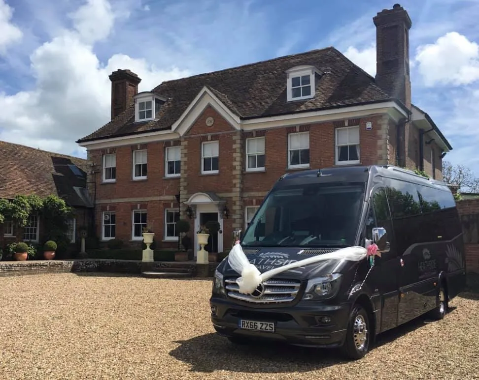 Silver Grey 19-seater Mercedes Sprinter in front of wedding venue decorated with large fluffy white ribbons and pink bows on its wing mirror