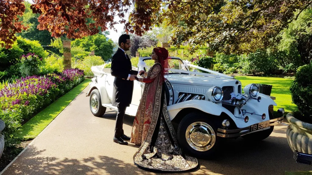 White Convertible Beauford Car with Roof open in the middle of a park with plenty of flowers. Asian Bride and her groom standing in front of the vehicle looking at each other while holding hands