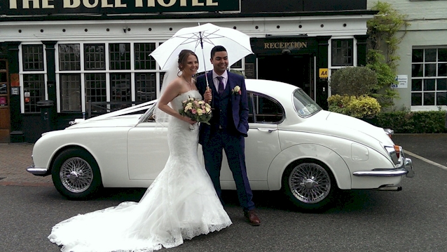 Bride and Groom in front of a Classic Jaguar. Bride is wearing a white wedding dress white the groom is holding a white umbrella in his right hand