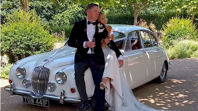 Bride and Groom posing for photos in front of a Classic Jaguar Mk2. Grooms wear a black suit with bow tie and the Bride wears a white dress holding the groom's hand