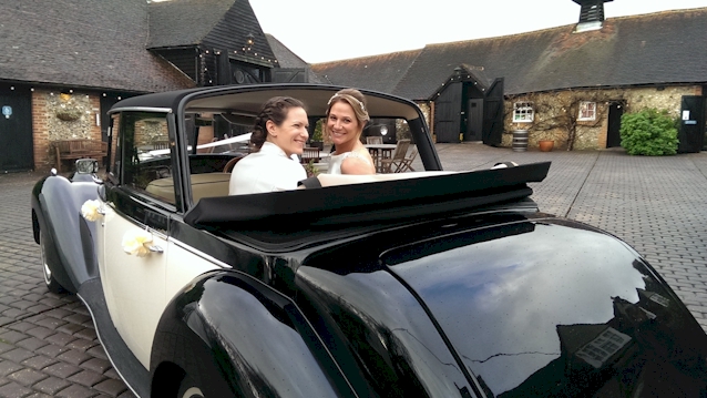 Two Brides in the rear of a Vintage Convertible Royale Wedding car with Roof Down looking backward at their photographer.