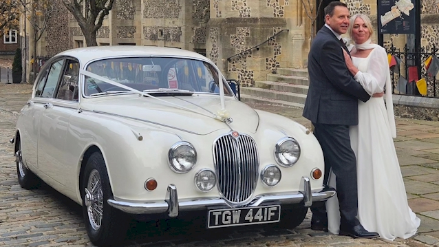 Front view of Mk2 Jaguar with pale pink Ribbons accross the front of the vehicle. Both Bride and Groom are standing next to the car posing for their photographer