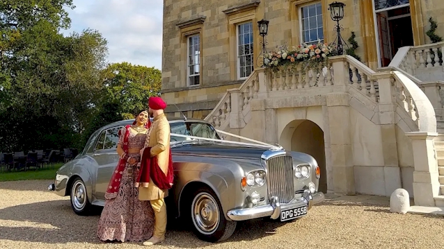 Classic Bentley S3 in front of wedding venue with Asian Bride and Groom in Red and Gold Wedding outfits standing in front of the vehicle looking at each others