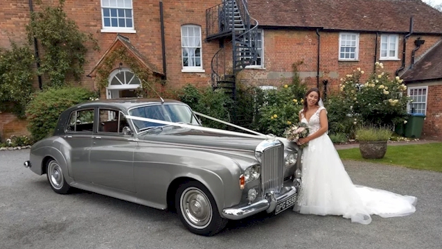 Right side view of a Silver Bentley S3 with Bride wearing a white dress and holding a bouquet of flowers stood in front of the vehicle
