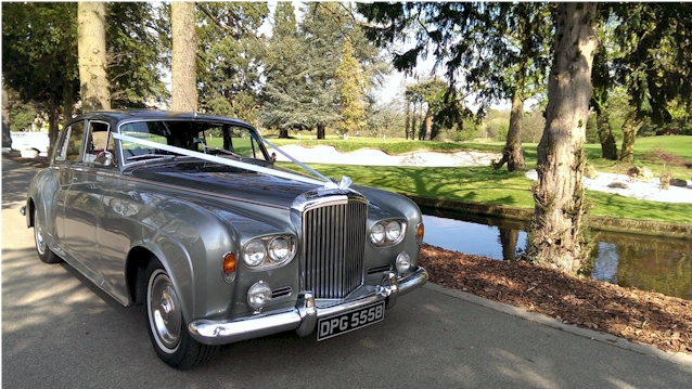 Front side view of a classic silver Bentley S3 with white wedding ribbons.