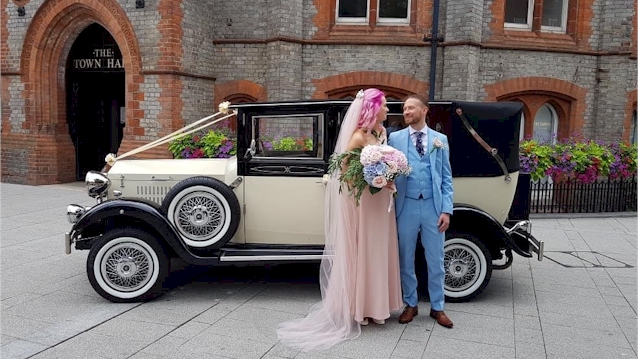 Bride and Groom standing in front of vintage car/ Bride is wearing a light pink wedding dress and groom a light blue suit