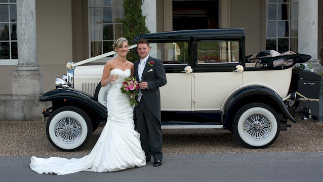 Bride and groom standing in front of a Black & White Vintage Badsworth convertible with roof down in front of a wedding venue
