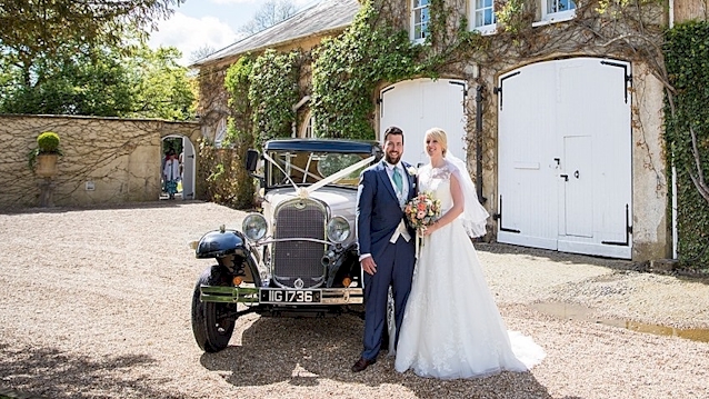 Front viw of Vintgae car with bride and grom standing in front of the vehicle for photo
