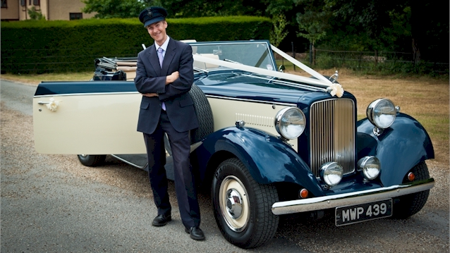 Fully uniformed chauffeur with grey suit and hat standing in front of the vintage royale convertible car