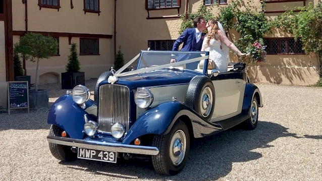 front view of vintage car with roof down. Bride and groom standing in the back of the vehicle kissing
