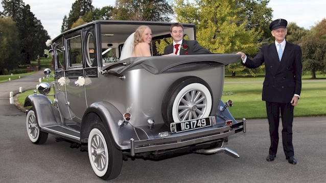Rear view of the Silver Imperial with roof down. Bride and Groom are inside the vehicle looking backward and theie chauffeur standing next to the vehicle with his chauffeur's hat