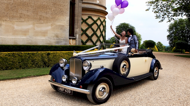 Left front view of vintage convertible car with white wedding ribbons and Brideand Groom standing in the rear of the vehicle holding some pink and purple ballons