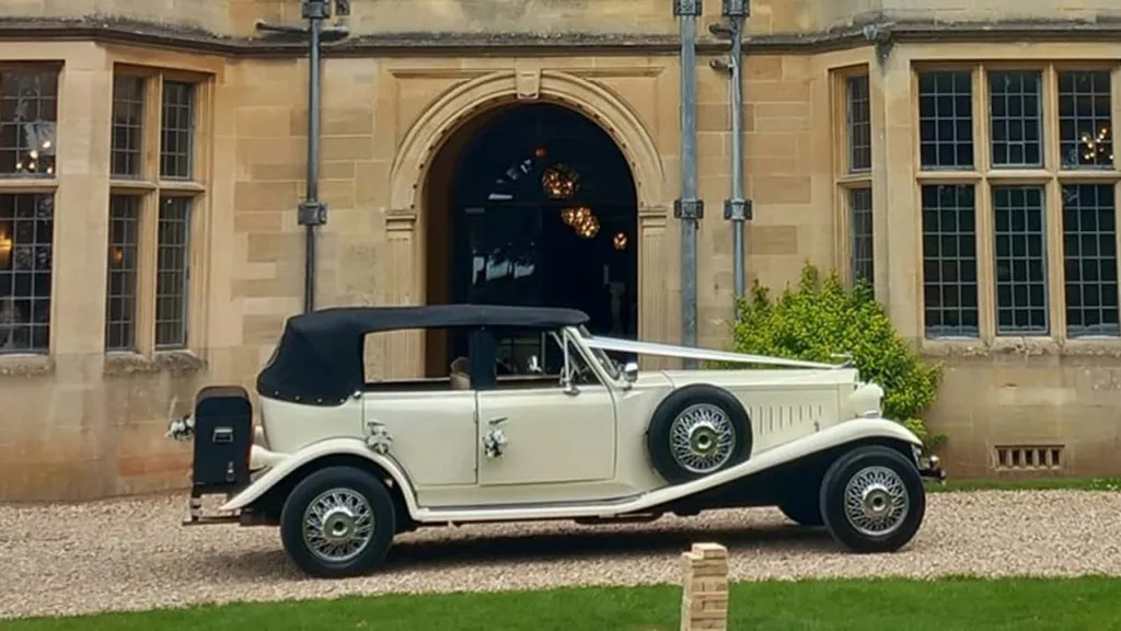 Right Side view of Ivory Beauford with Black Roof up, Spare wheel mounted on right wheel arch and Traditional V-shape white ribbon accross the bonnet in front of wedding venue
