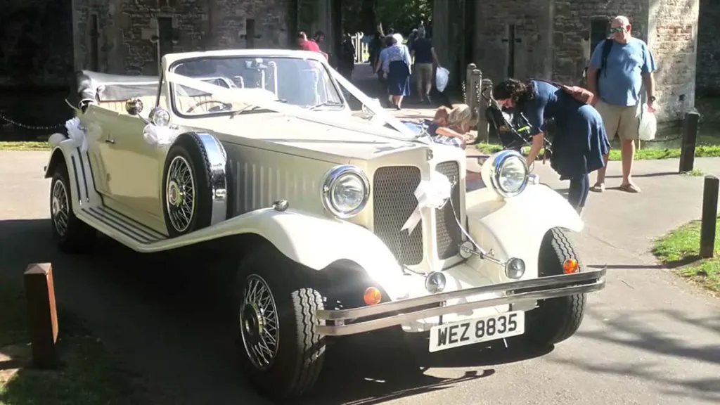 Full front view of a Vintage Beaford with roof down and ribbon decoration at the front in attendance at a wedding. Some wedding guests are standing in the background