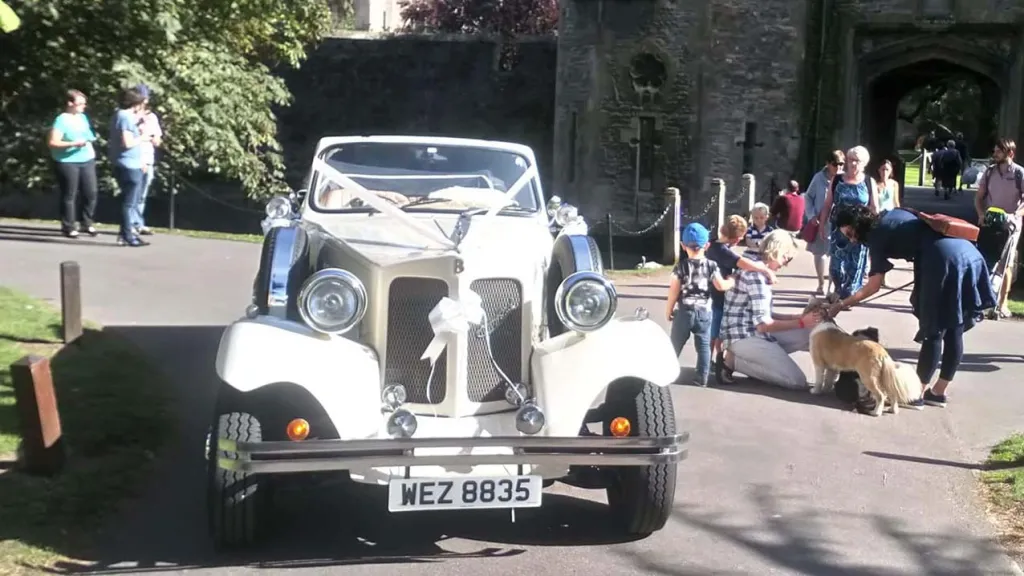 Full front view of a Vintage Beaford with roof down and ribbon decoration at the front in attendance at a wedding. Some wedding guests are standing on the side of the vehicle