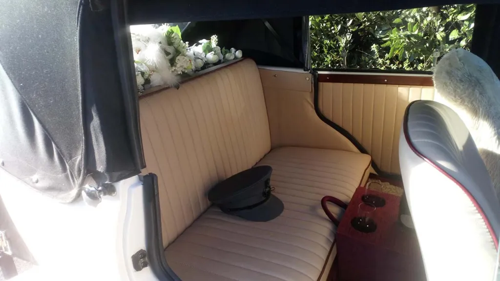 Inside rear bench seat in cream leather with artificial white flowers and green foliage on parcel shelf, chauffeur's hat on the seat and rear passenger door is open showing large legroom