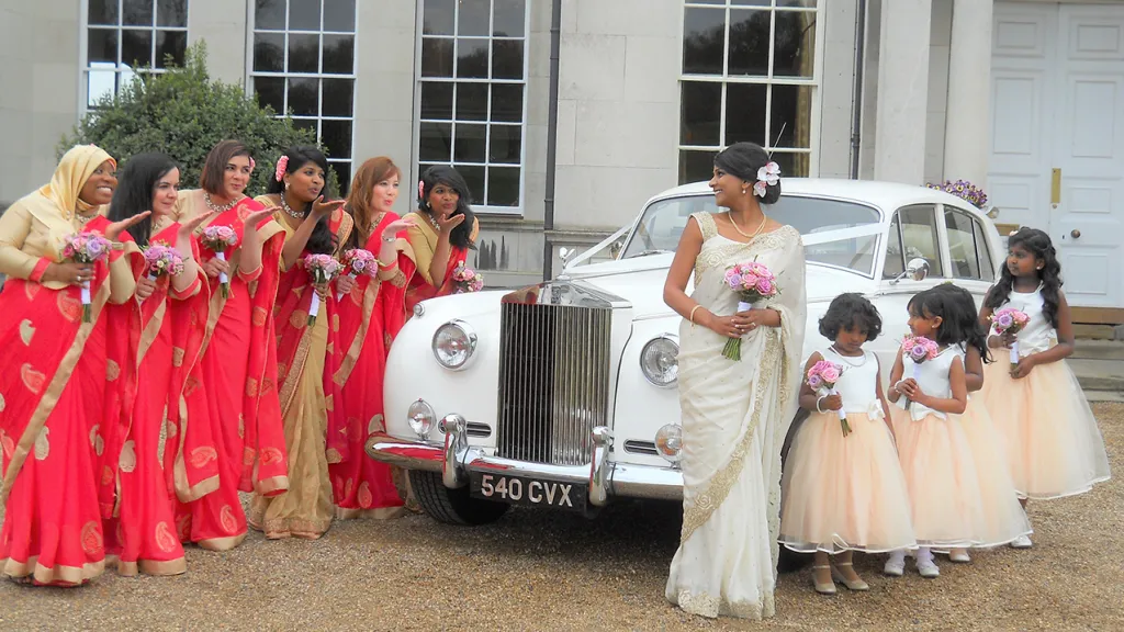 Rolls-Royce Silver Cloud in the Middle of bridesmaids wearing red dressed and flower girls in Ivory dress
