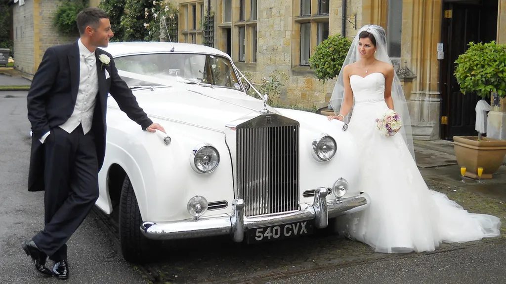 Bride and Groom leaning on the bonnet of a Classic White Rolls-Royce. Bride is wearing a white wedding dress and holds a bouquet of flowers in her left hand
