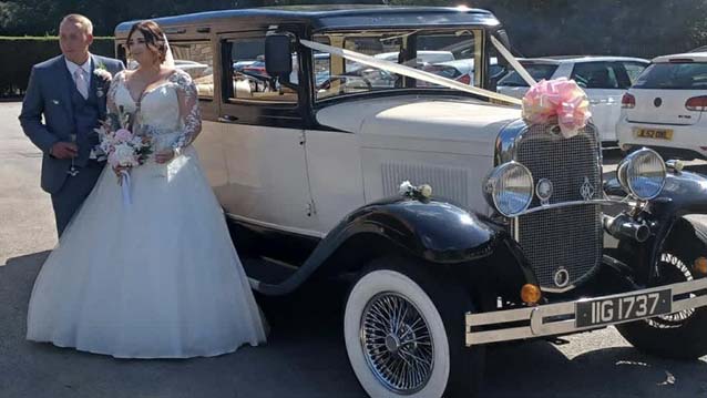 Vintage Bramwith decorated with large pale pink bow on top of front grill with matching Ribbons accros its bonnet. Both Bride and Groom standing in front of the vehicle for photos