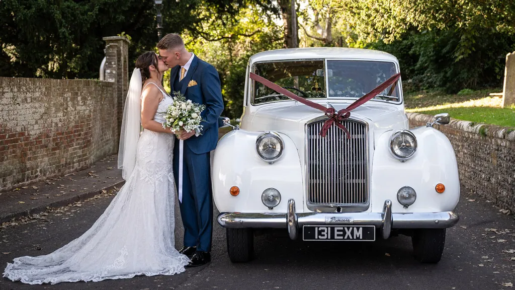Bride and Groom standing and kissing next to a Classic Austin Princess in Ivory with burgundy wedding ribbons.