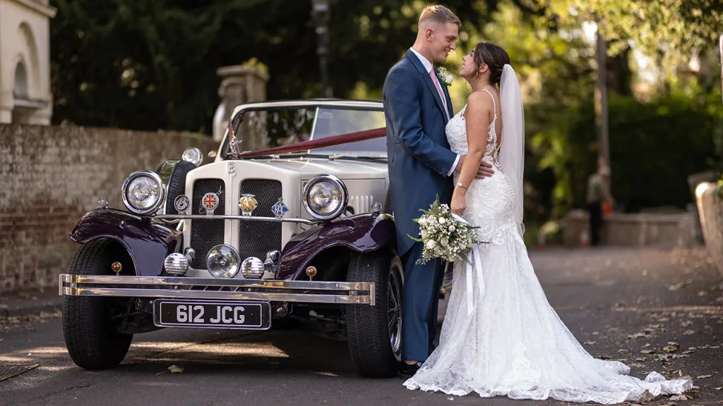 Bride and Groom holding hands oin front of a Convertible Vintage Car dressed for with burgundy wedding ribbons