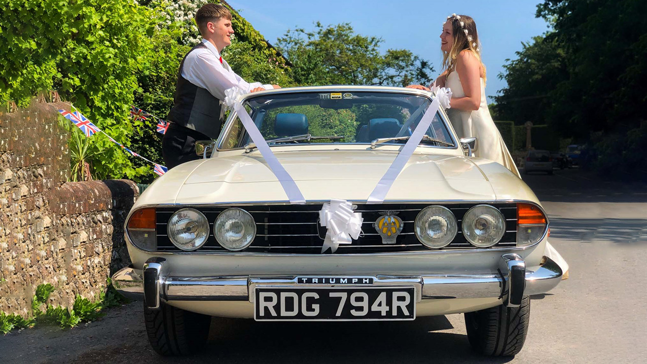 Full front view of Classic Triumph Stag Convertible dressed with white ribbons and large bow on the front grill. Bride and Groom are standing on either side of the vehicle