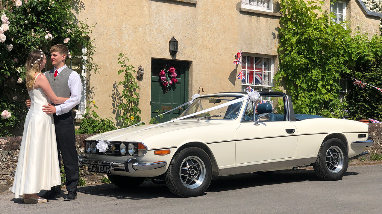 Bride and Groom holding each other in front of a Ivory Triumph Stag Convertible