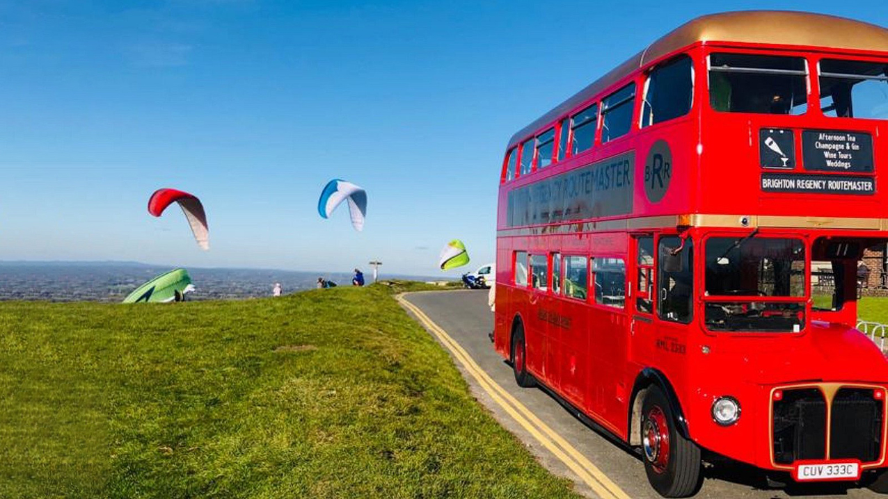 Double decker red routemaster on top of a hill with view of the sea in the background and some paruchutists in the blue sky