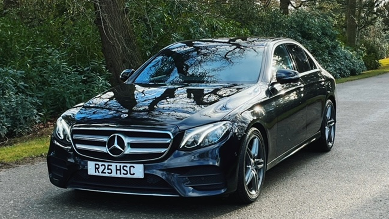 Front view of Black Mercedes on tarmac with green trees in the background