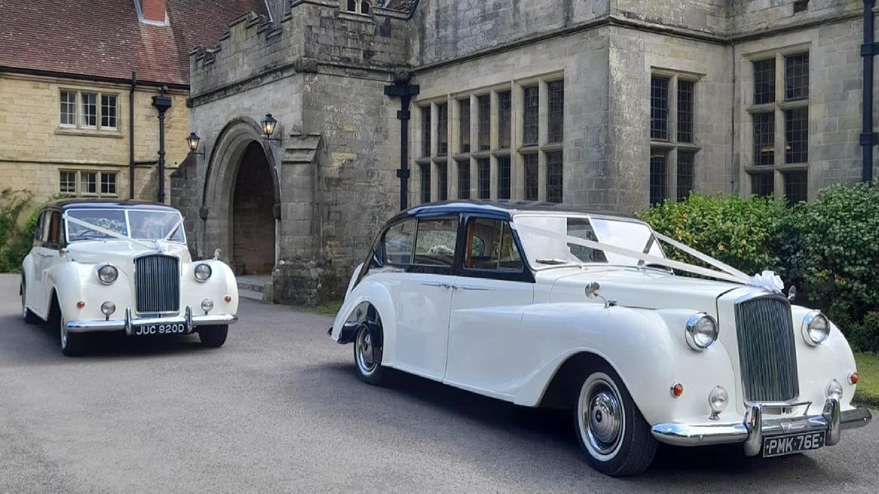 Two Classic Austin wedding cars decorated with white ribbons and bow accross front bonnet in front of wedding venue near Brighton