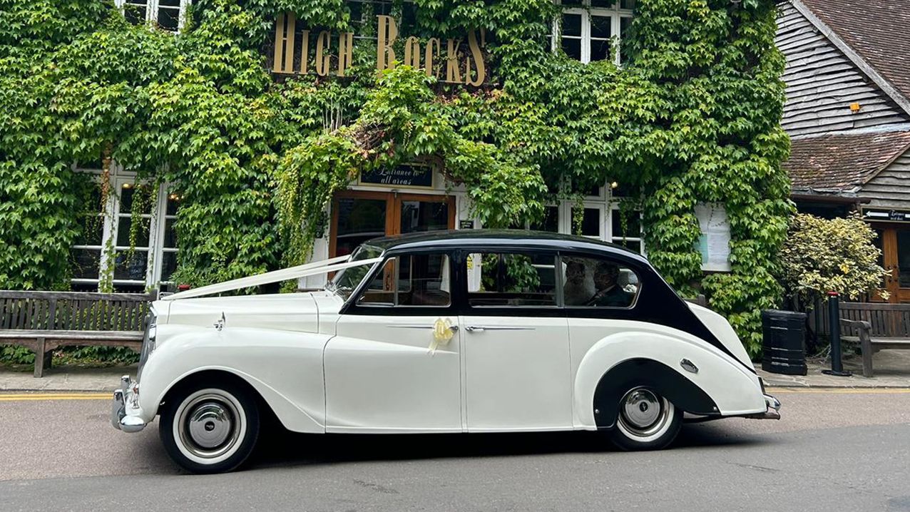 Left view of Classic Austin Princess decorated with ivory ribbons. Background is a wedding venue covered with green ivy.