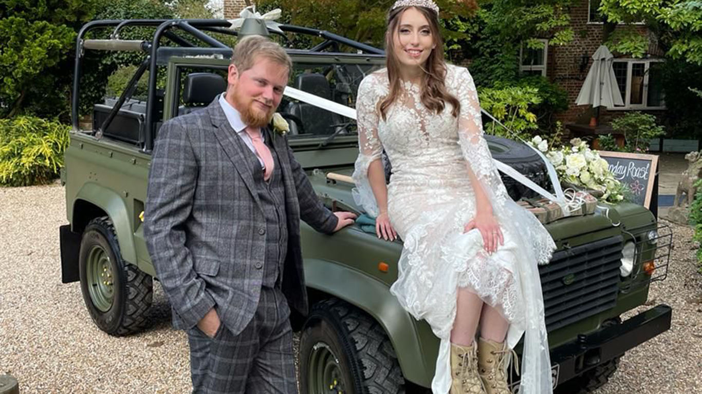 Bride wearting a white dressed seating on the bonnet of a Military Landrover decorated with ribbons and flowers. Groom standing by the vehicle