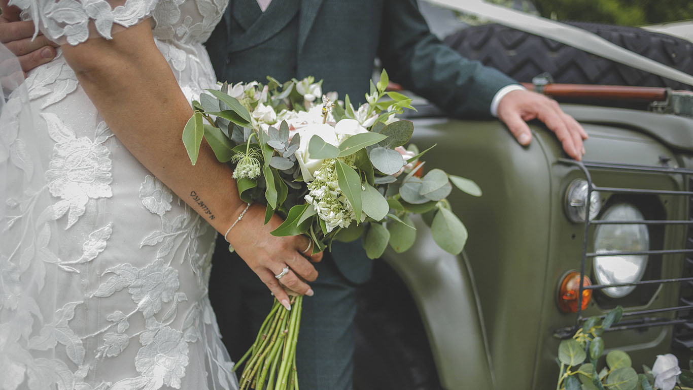 Bride holding a Green and White Bridal Bouquet next to the Groom and Landrover Car.