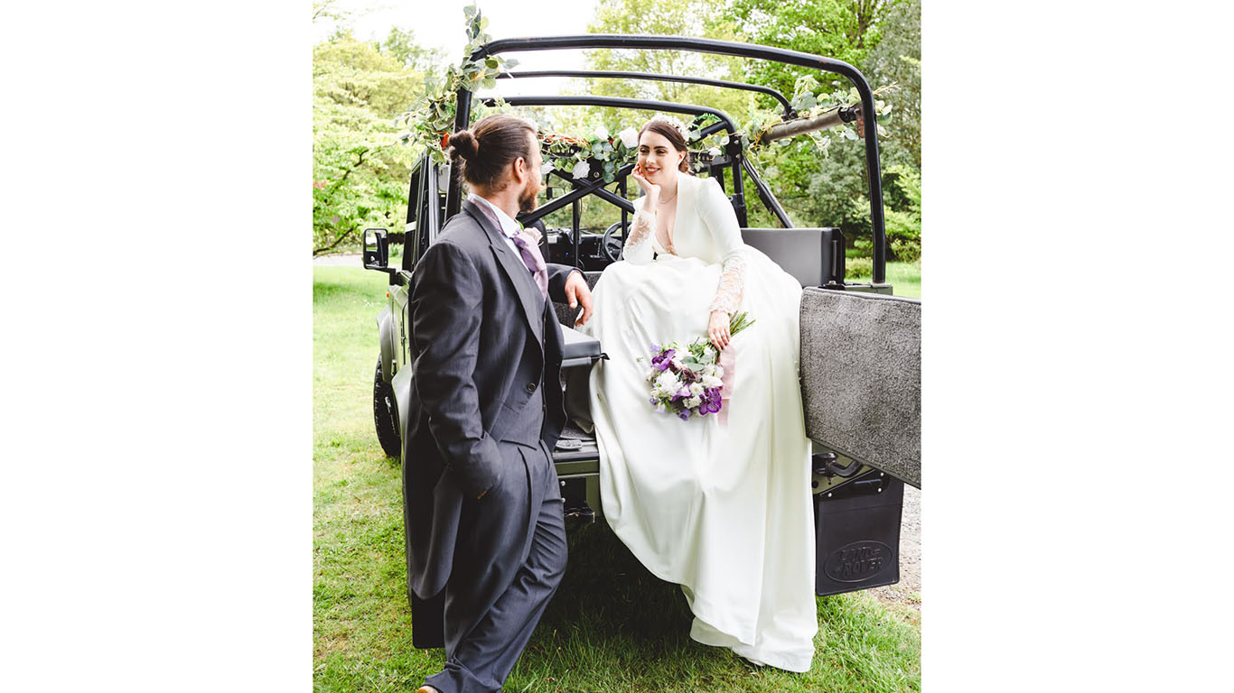 Bride wearing a large white wedding gown seating in rear seats holding a bridal bouquet looking at her groom standing by the vehicle