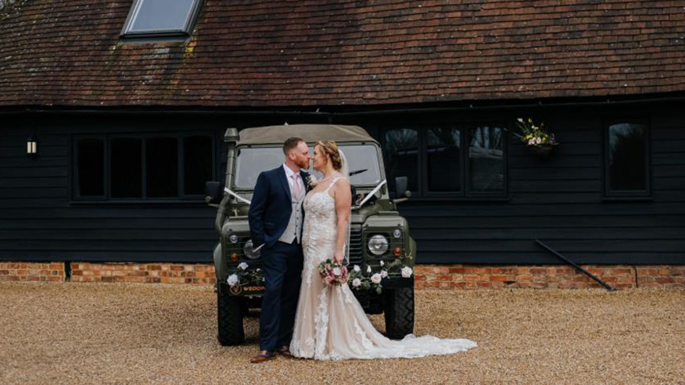 Bride and Groom kissing in front of Landrover decorated for wedding with ribbons and flowers in front of a wedding barn in Kent