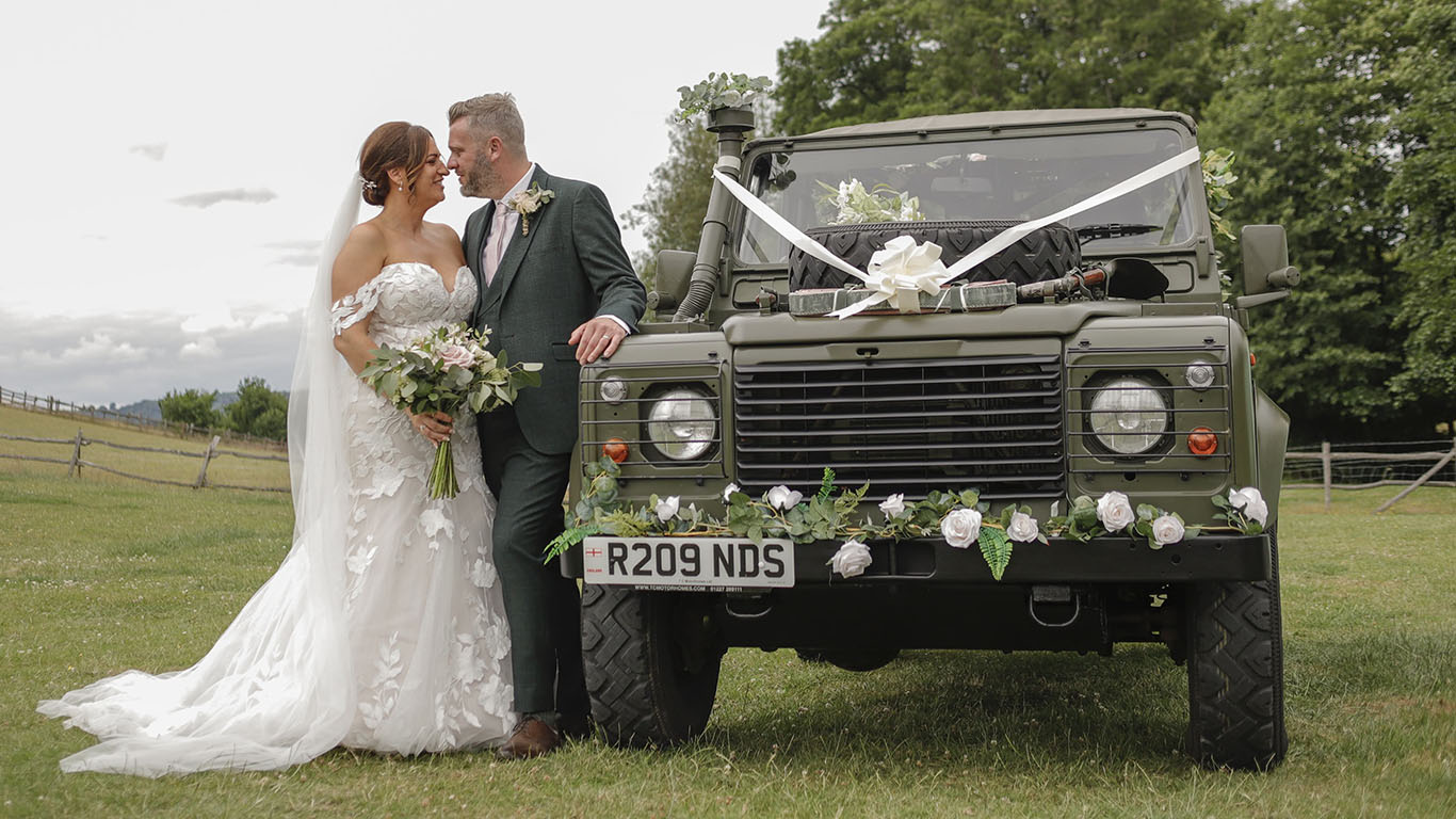 Bride and Groom standing on the side of the Landrover Military Vehicle dressed with floral decoration on front bumper and White ribbons and Bows on top of its bonnet.