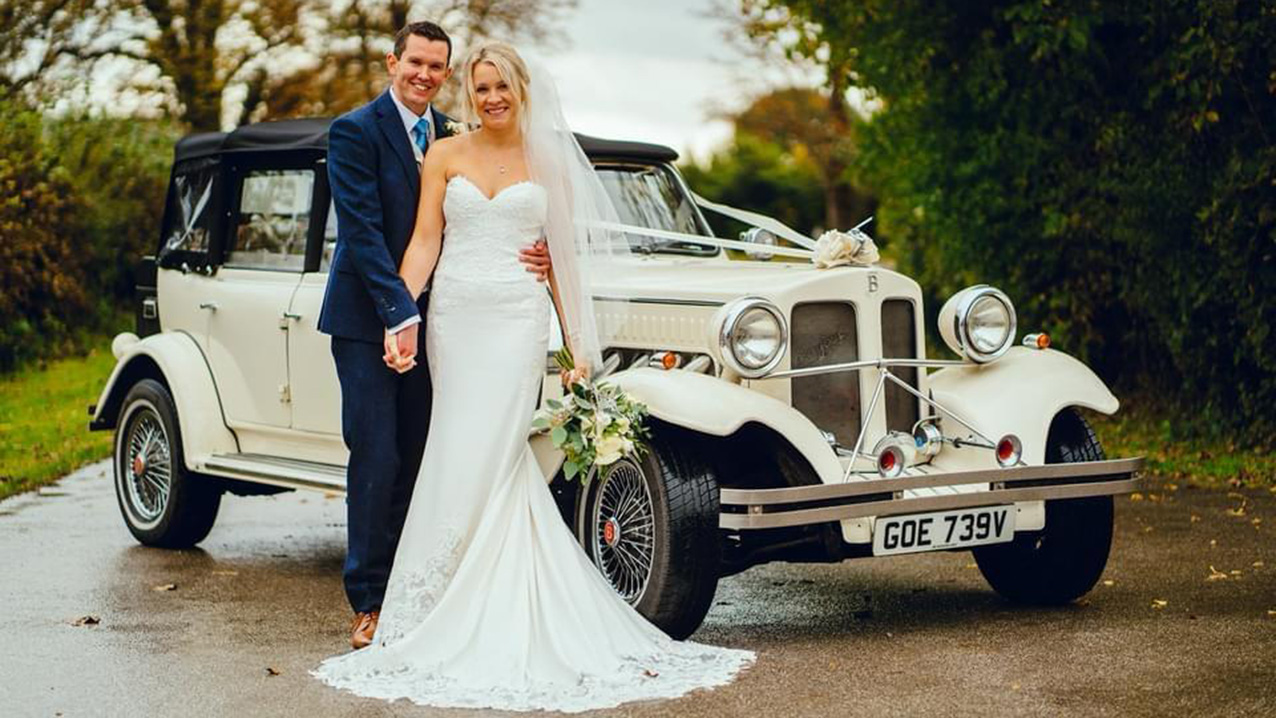 Bride and Groom standing in front of a Vintage Beauford in a path with Green Trees on either side