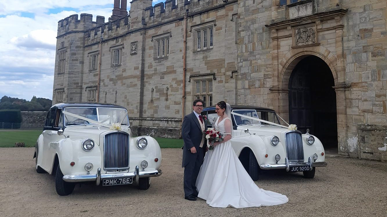Bride and Groom standing between two Austin Princess Limousine decorated with white wedding ribbons in front of a wedding castle in Sussex