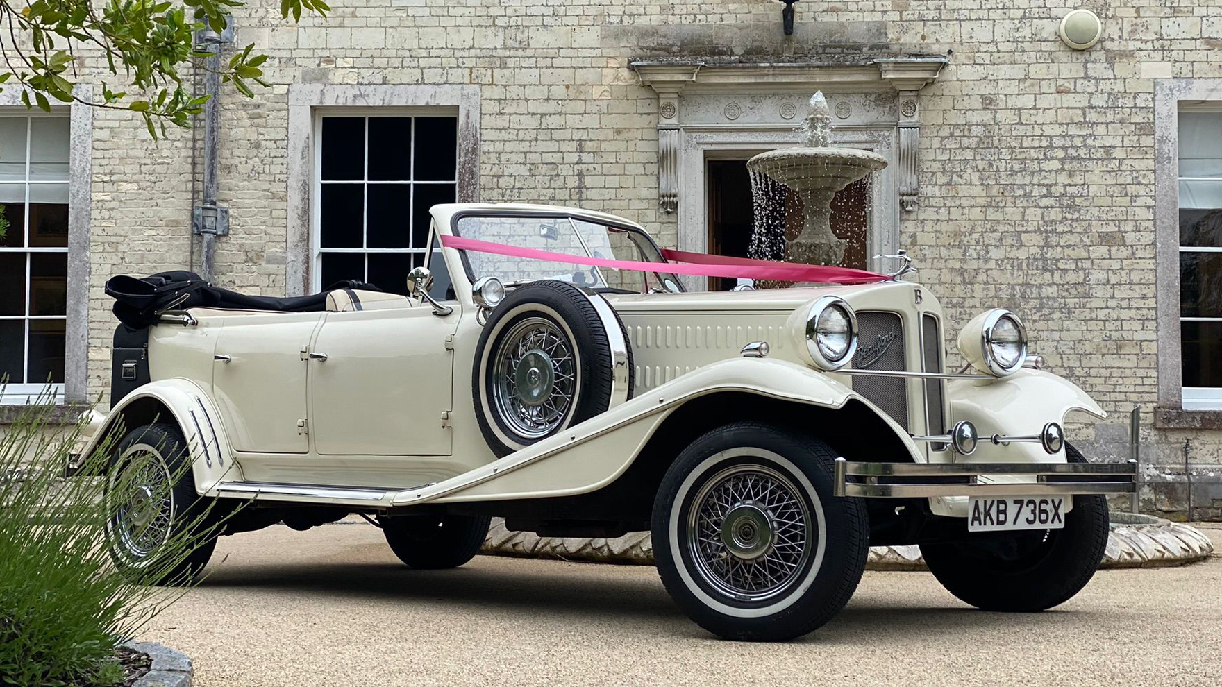 Front Right side of BEauford with Convertible Roof down decorated with Red Ribbons accross its bonnet. Wedding Venue shown in the back ground