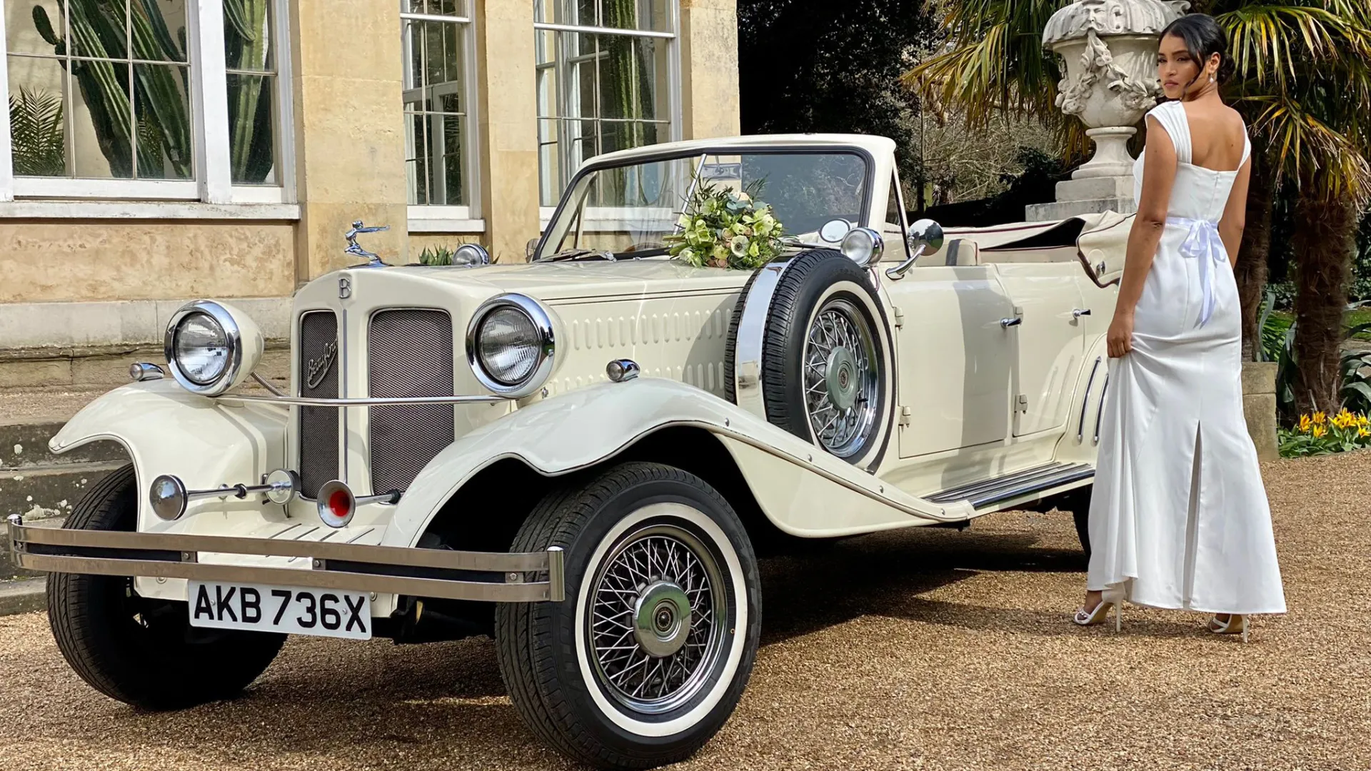 Front Side View of Beauford with convertible roof down with wedding flowers on top of the bonnet. Bride is standing by the vehicle posing in her white dress