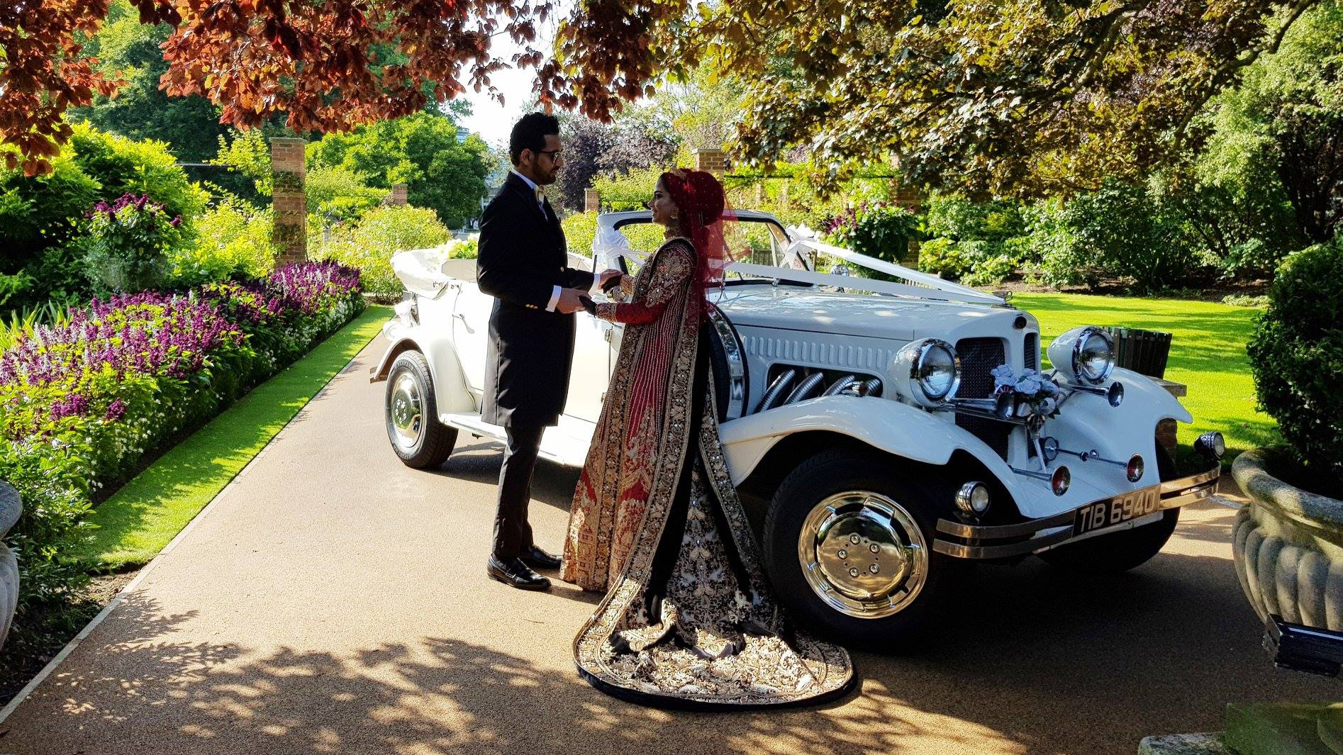 White Beauford convertible with roof down decorated with white ribbon in a park. Asian Bride standing in front of the vehicle with her groom