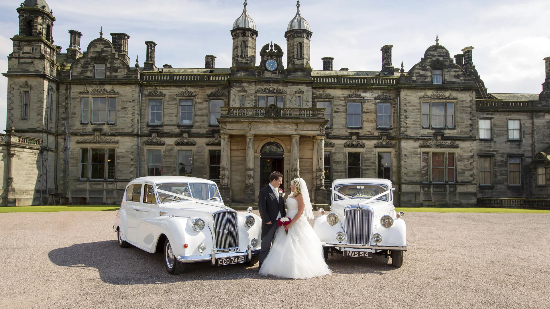 Two Classic Austin Limosuine decorated with matching Wedding Ribbons with bride and groom standing in the middle of the two vehicles