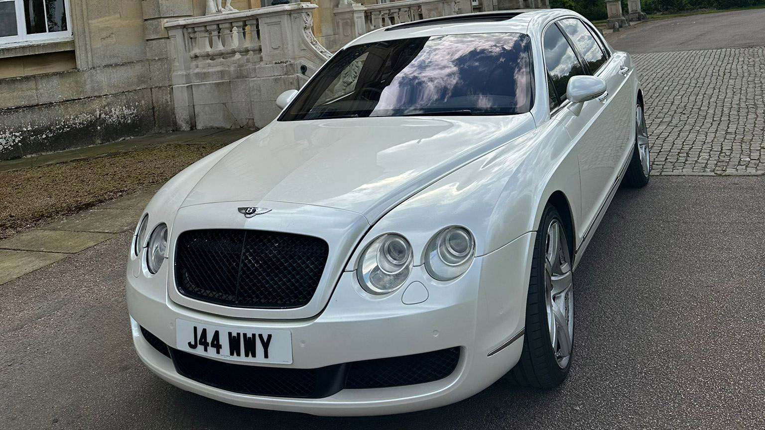 Front view of a Modern White Bentley with large Front Grill in Black in front ot a brick wall
