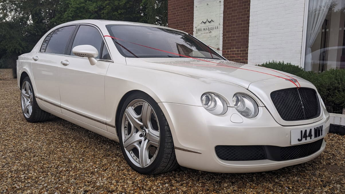 Front Right Side view of White Bentley Flying Spur showing the Chrome Alloy Wheels and double twin headlights in front of a wedding venue in Bedford