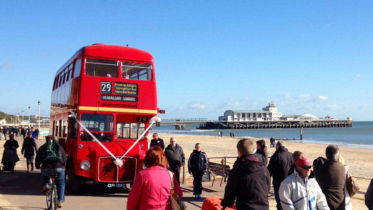 Red Double Decker Bus on Dorset Coastline with view of the sea in the background and wedding guests loading onto the bus