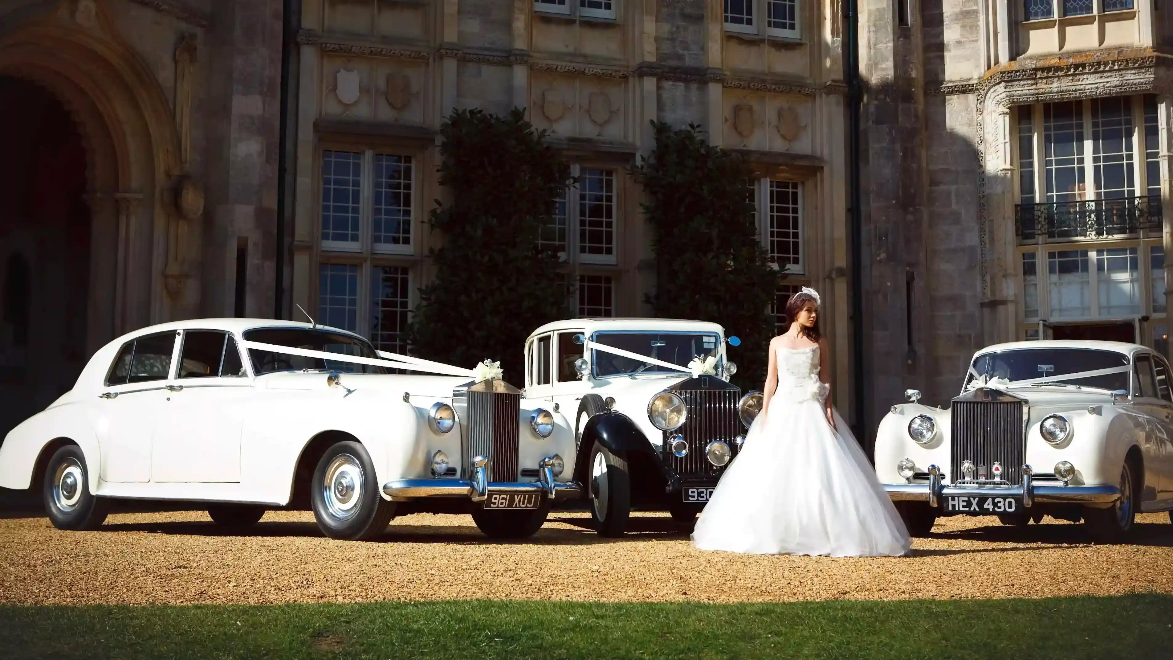 Selection of three Classic and Vintage Wedding Cars in White decorated with matching white ribbons and Bride wearing a white wedding dress standing in front of the vehicles.
