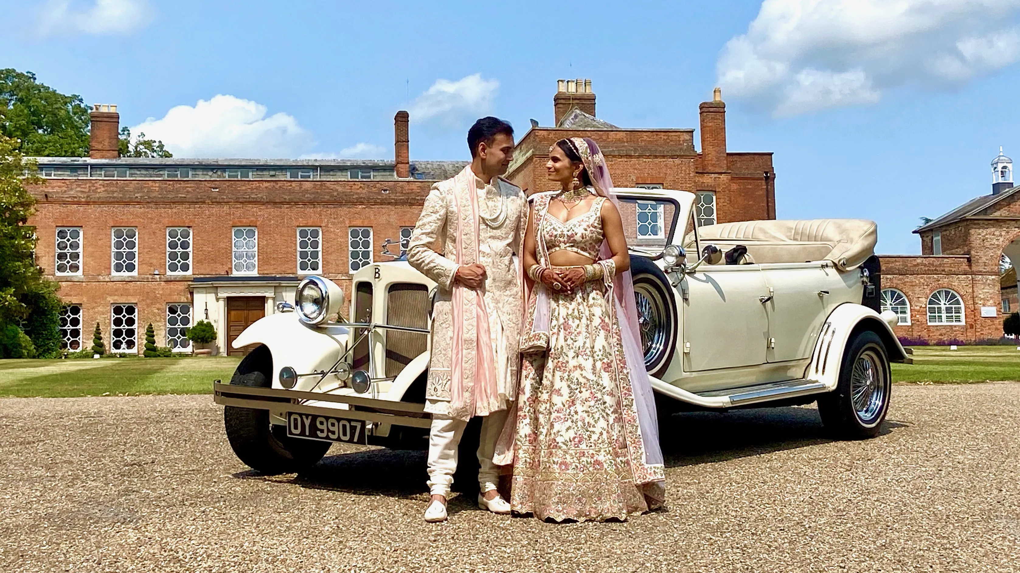 Convertible vintage style Beauford with roof down with newly wed Asian couple standing in front of the vehicle looking at each others. Vehicle is parked in front of a large manor house in Welling