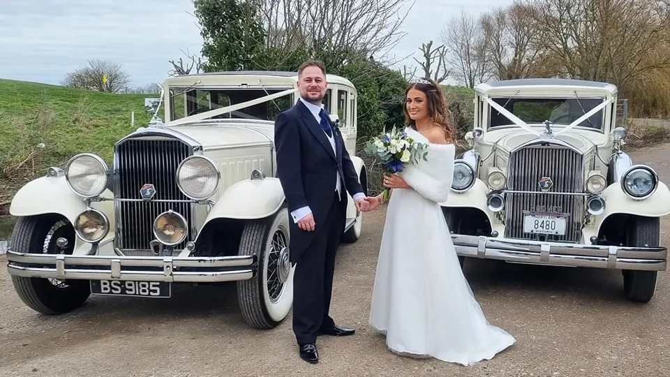 Two Ivory vintage wedding cars decorated with ivory ribbon at a local Braintree wedding with Bride and Groom standing in the middle of the cars holding hands.