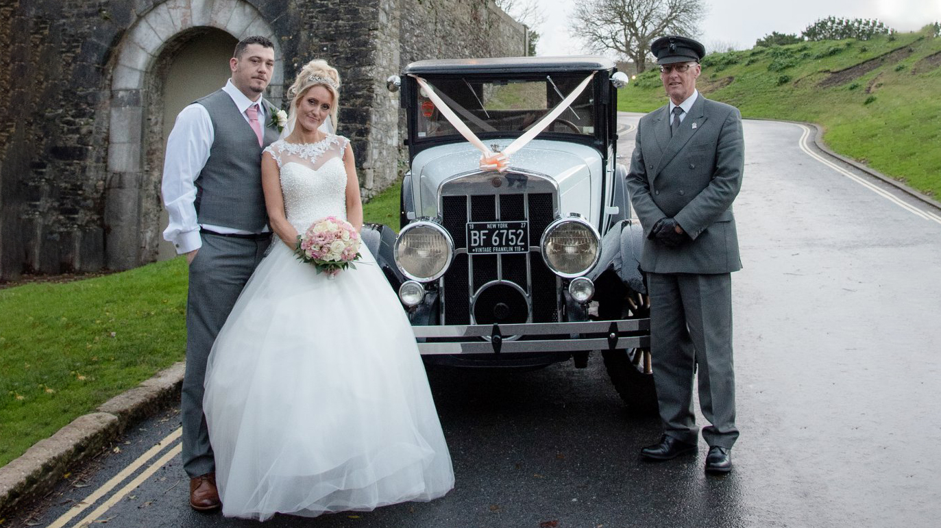 Bride and groom standing on the left with the Vintage Franklin car in the Middle and the Chauffeur wearing a grey suit and hat on the right