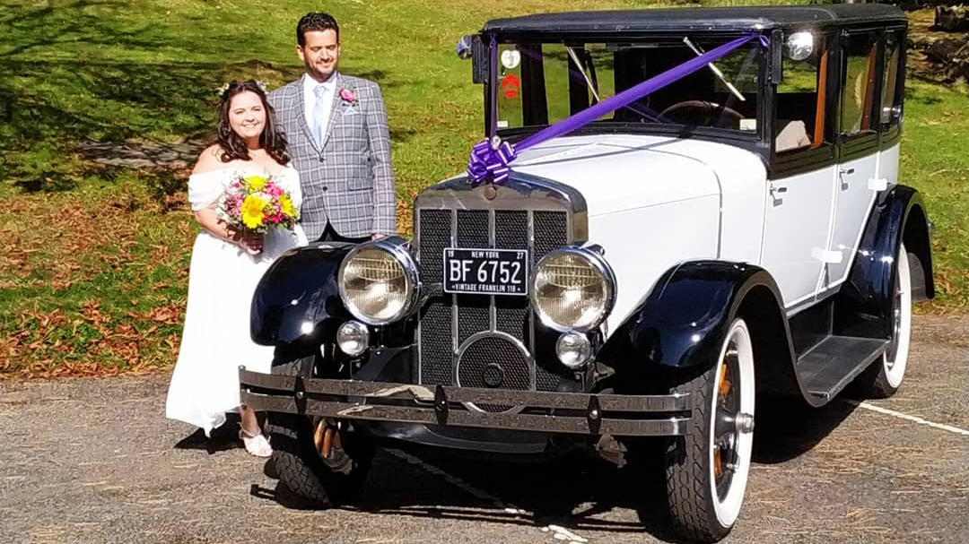 Bride and Groom standing on the side of the vintage car dressed with purple ribbons across the front bonnet.
